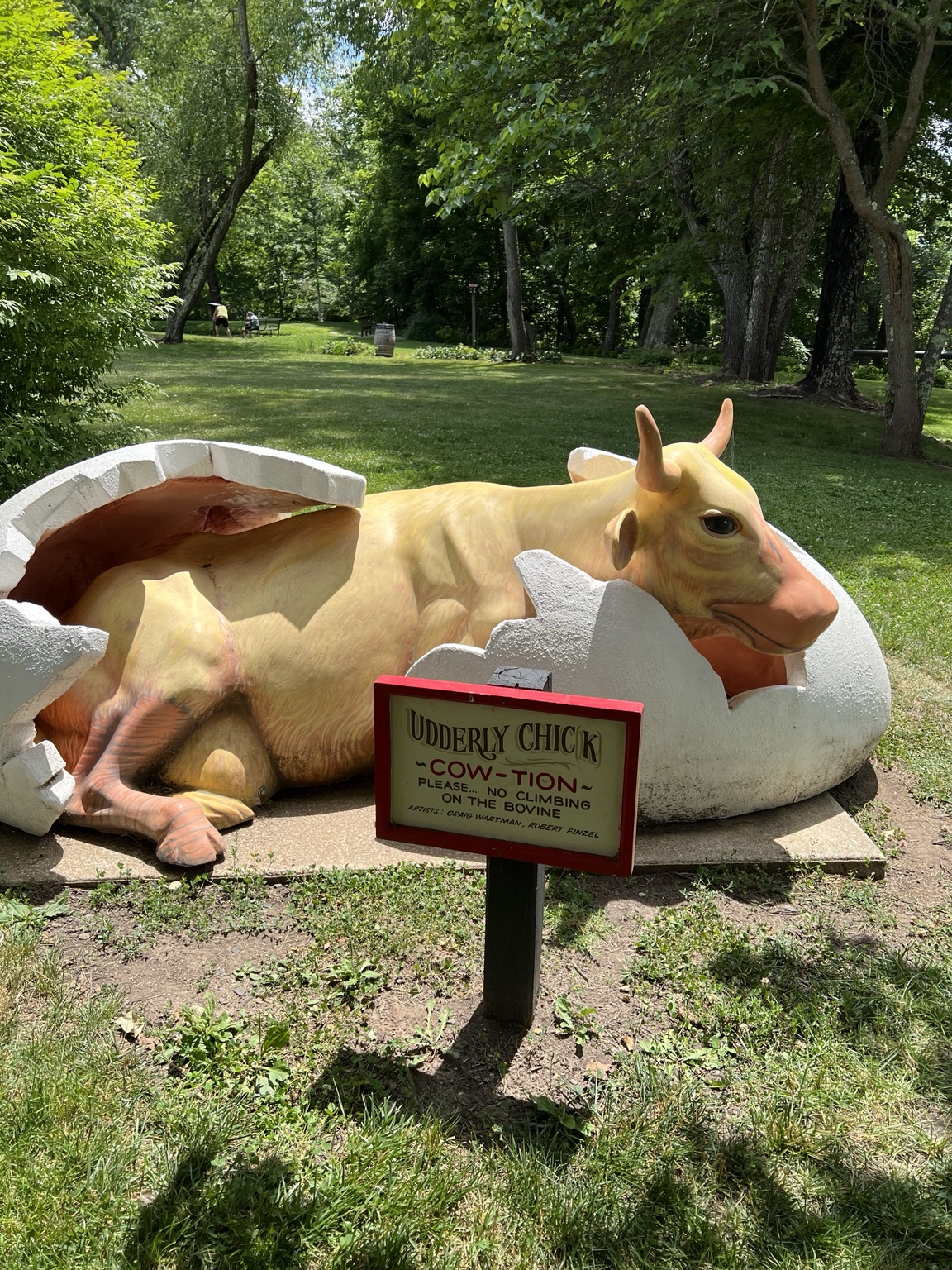 A whimsical painted cow sculpture emerging from a giant cracked eggshell on the lawn at Paint Bank, with a small “Udderly Chick” sign