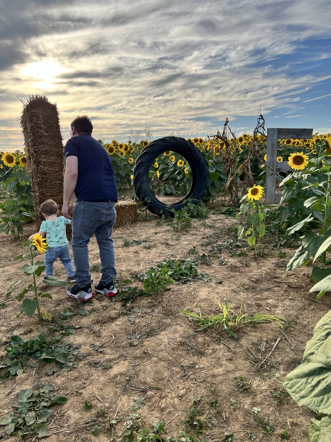 An adult and the Littlest Wayfinder walk over rough dirt and weeds toward a rustic tire photo setup