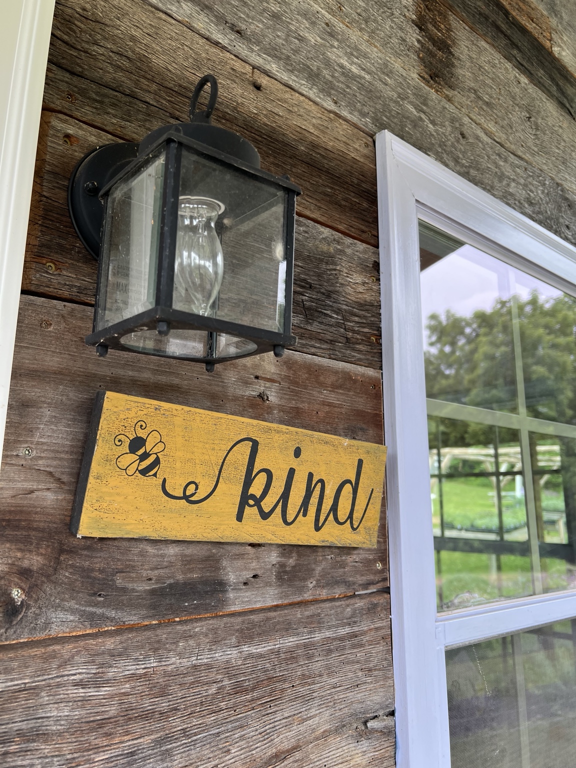 A yellow Bee Kind sign mounted next to a lantern and window on a rustic wooden building