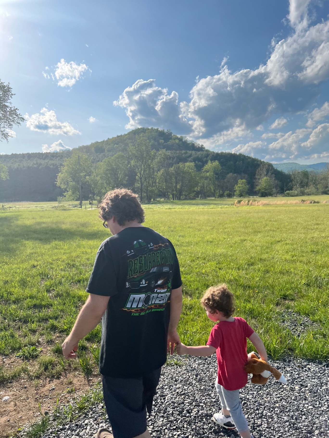 A father and young child walk hand in hand through an open field beneath the mountains at Hayfields State Park.