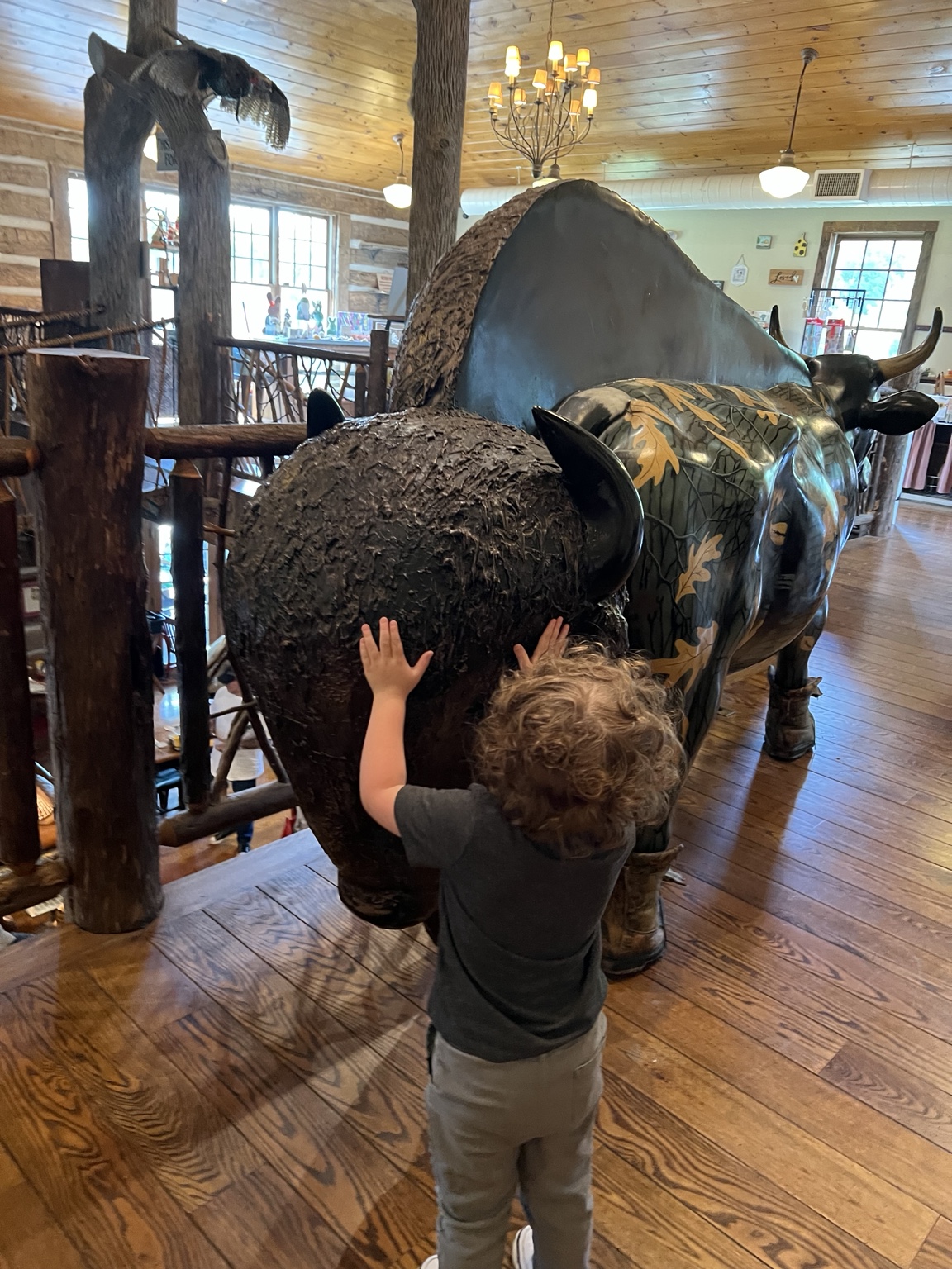A small child, seen from behind, reaches up to touch a life-sized painted buffalo sculpture inside the Paint Bank General Store