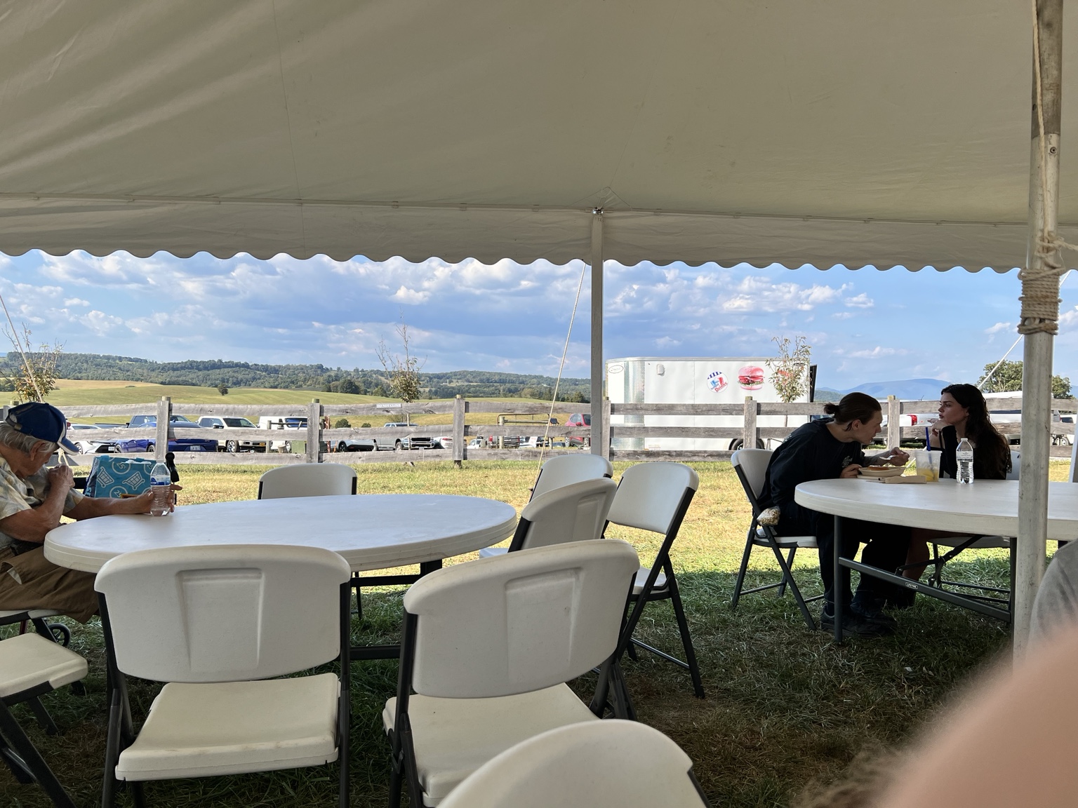 A covered communal dining tent with round tables and chairs, visitors eating, and parking and rural landscape visible beyond